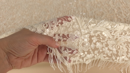 Colț de dantelă brodată cu micropaiete strălucitoare ținut în mână evidențiind franjurii de mărgele
