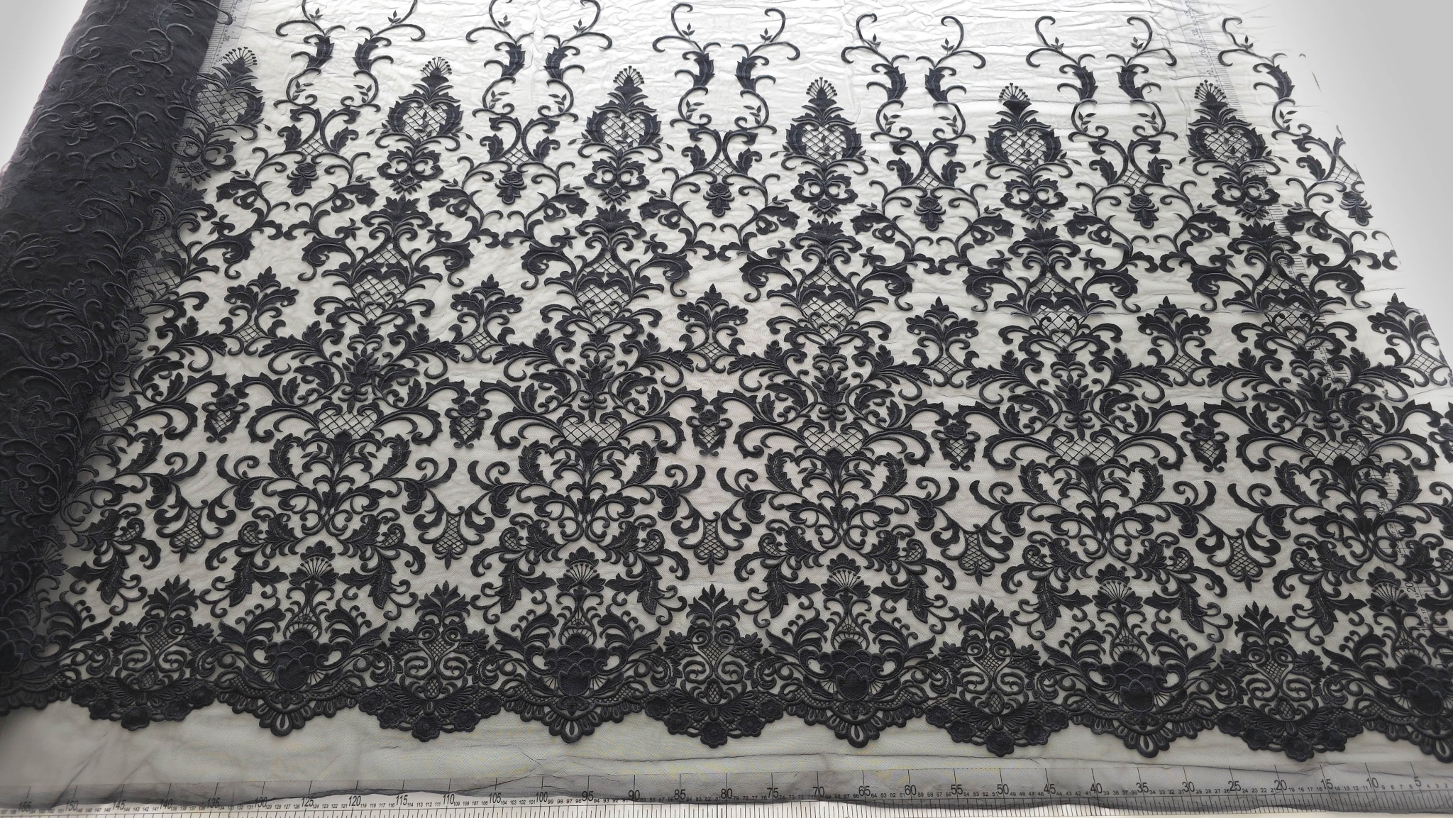 Dantelă neagră brodată pe tulle în stil baroc materialul desfășurat pe masă, cu bordură festonată vizibilă și riglă de măsurare în partea de jos