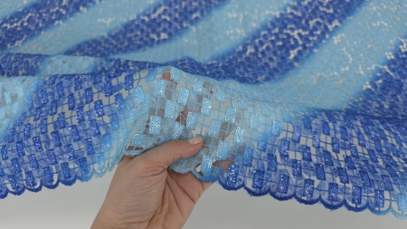 Mână ridicând bordura broderiei albastre degrade, evidențiind transparența între motive