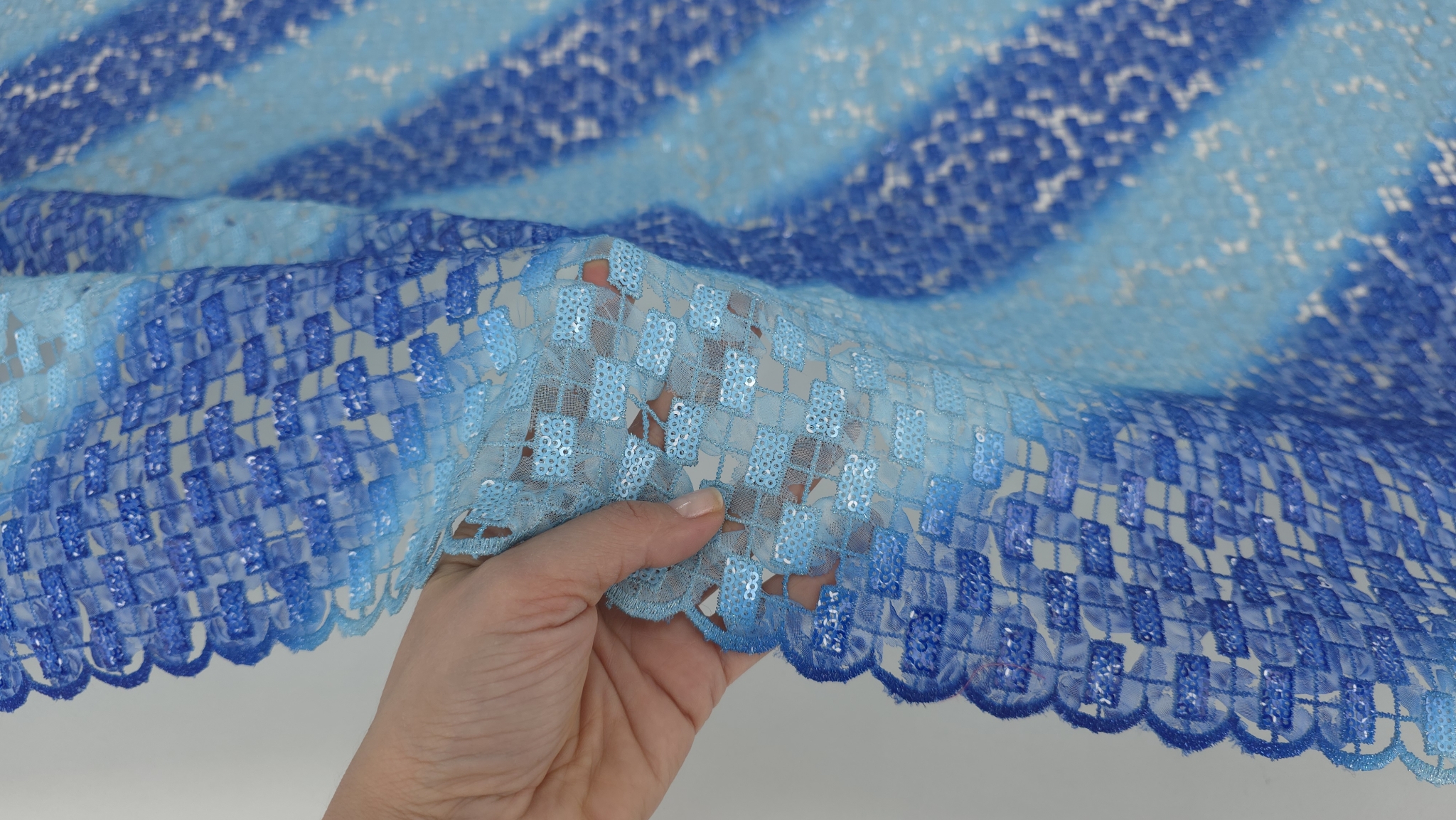 Mână ridicând bordura broderiei albastre degrade, evidențiind transparența între motive