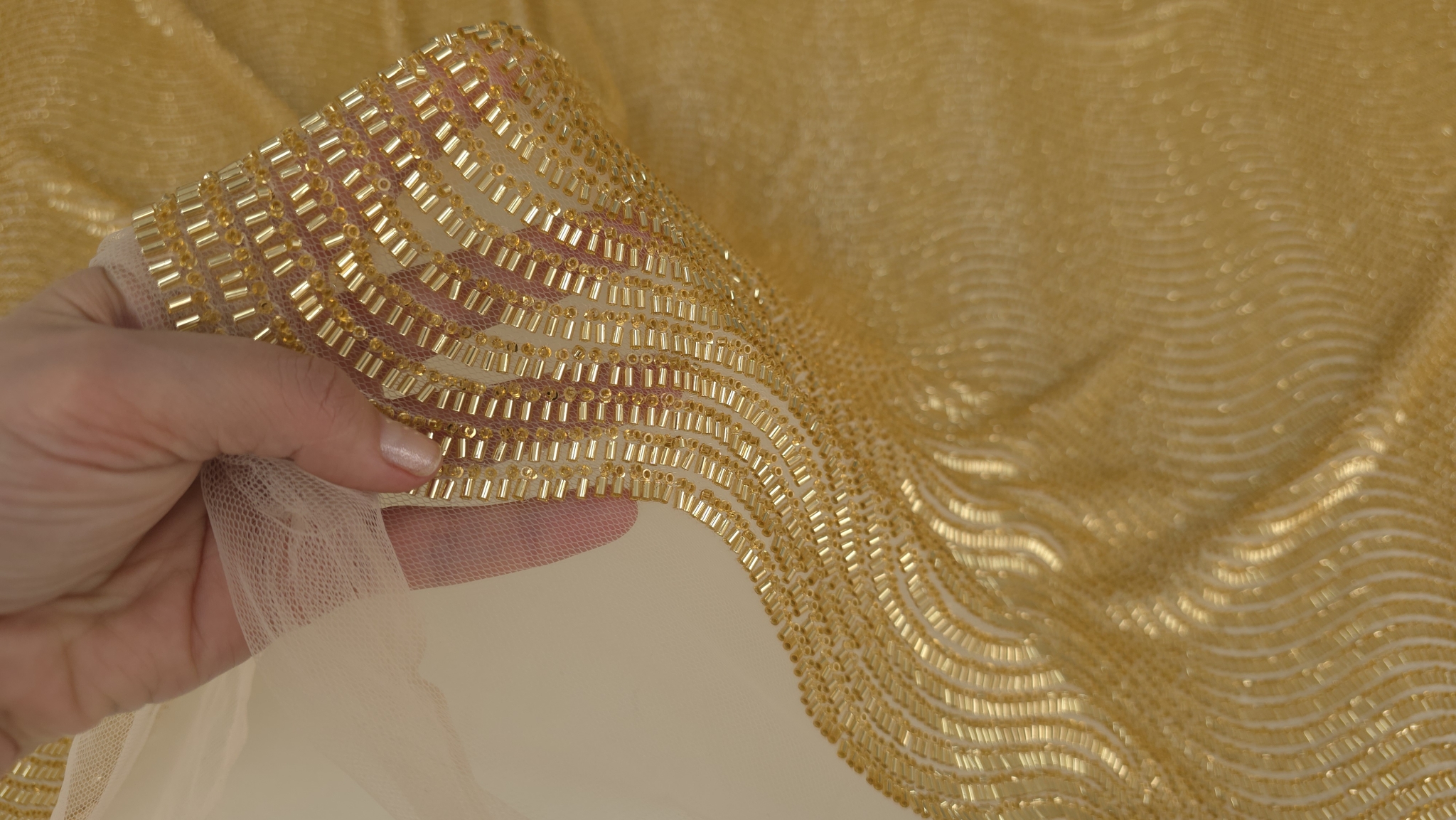 Mână ridicând marginea materialului auriu pentru a arăta tulul de bază și cusătura individuală a mărgelelor