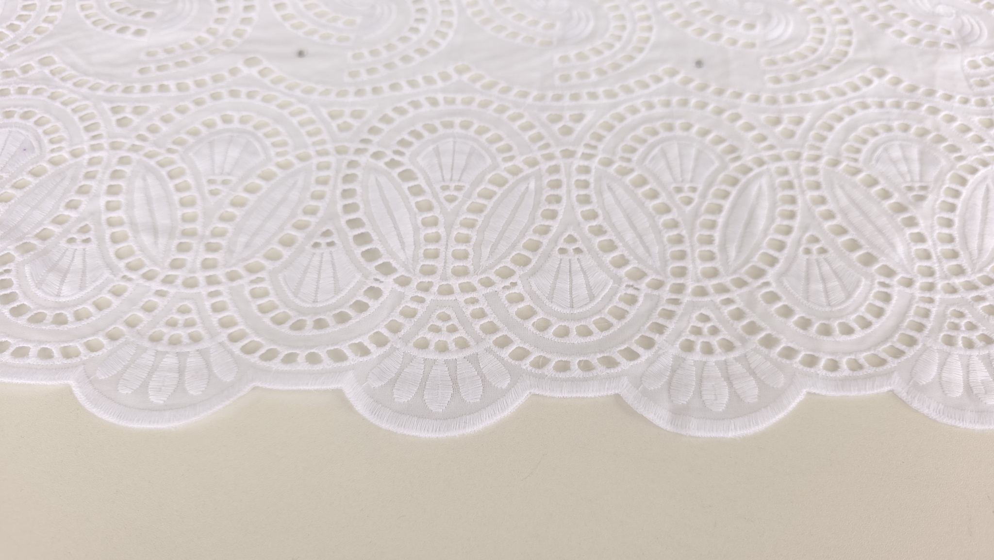 Suprafața dantelei brodate văzută de aproape, cu spirale și decupaje pătrate conturate de broderie albă