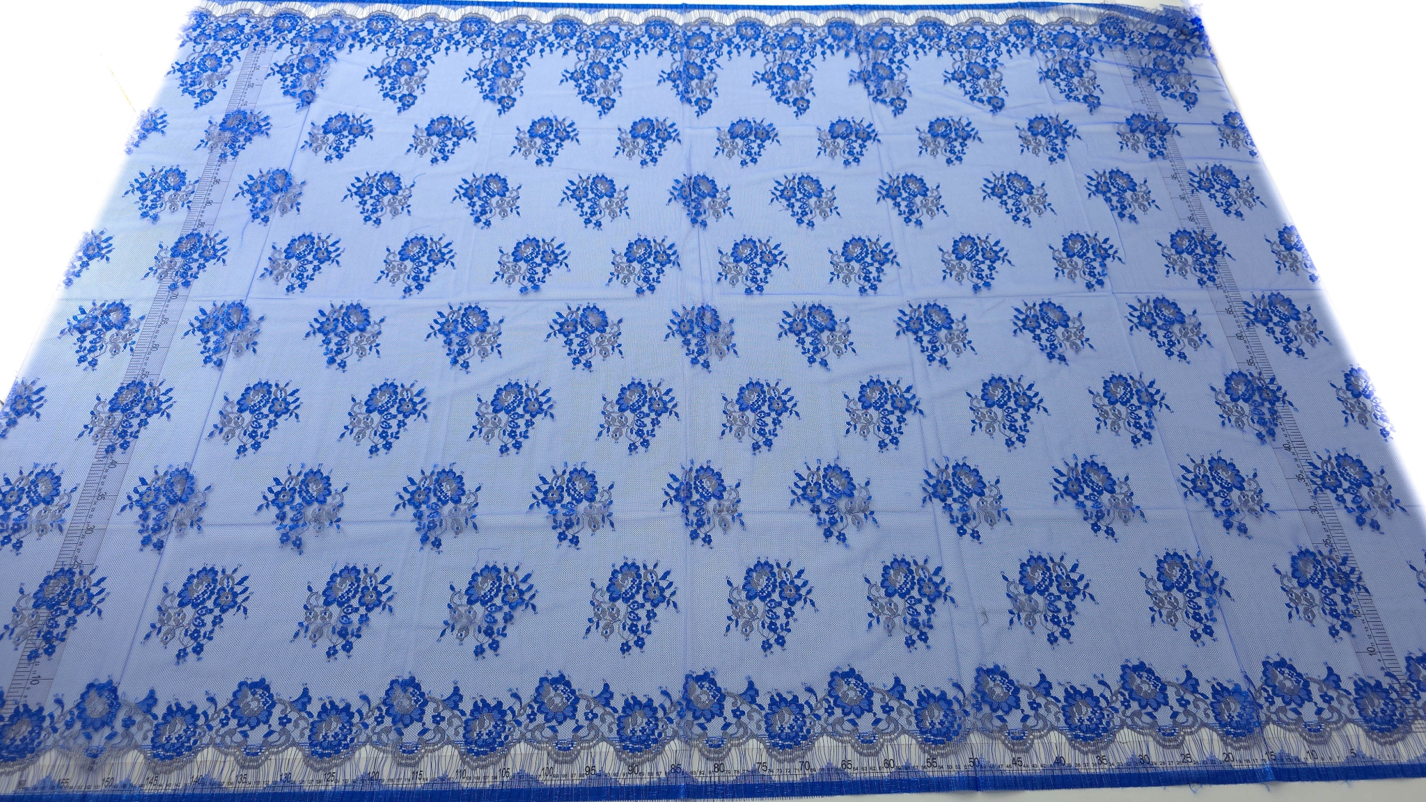 Dantela tip Chantilly albastru royal cu inserții argintii desfășurată pe masă cu rigle laterale și bordura dublă vizibilă