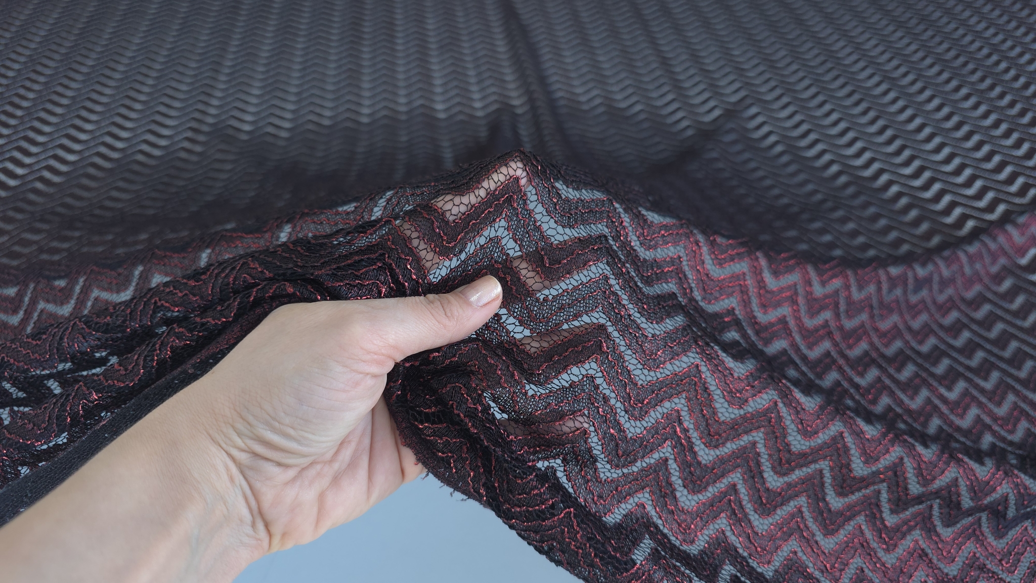 Detaliu al materialului ținut între degete cu reflexii bordeaux pe liniile zigzag