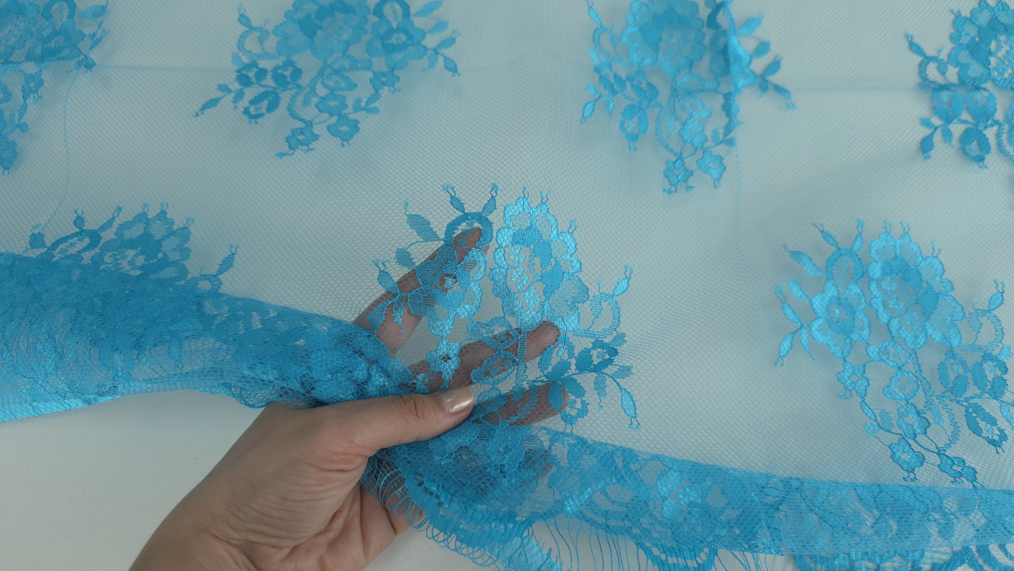 Mână care prinde bordura dantelei turquoise cu motiv floral vizibil prin tullul transparent pe palmă