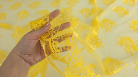 Mână care ridică bordura galbenă cu franjuri și motive florale dense pe fond de tull transparent