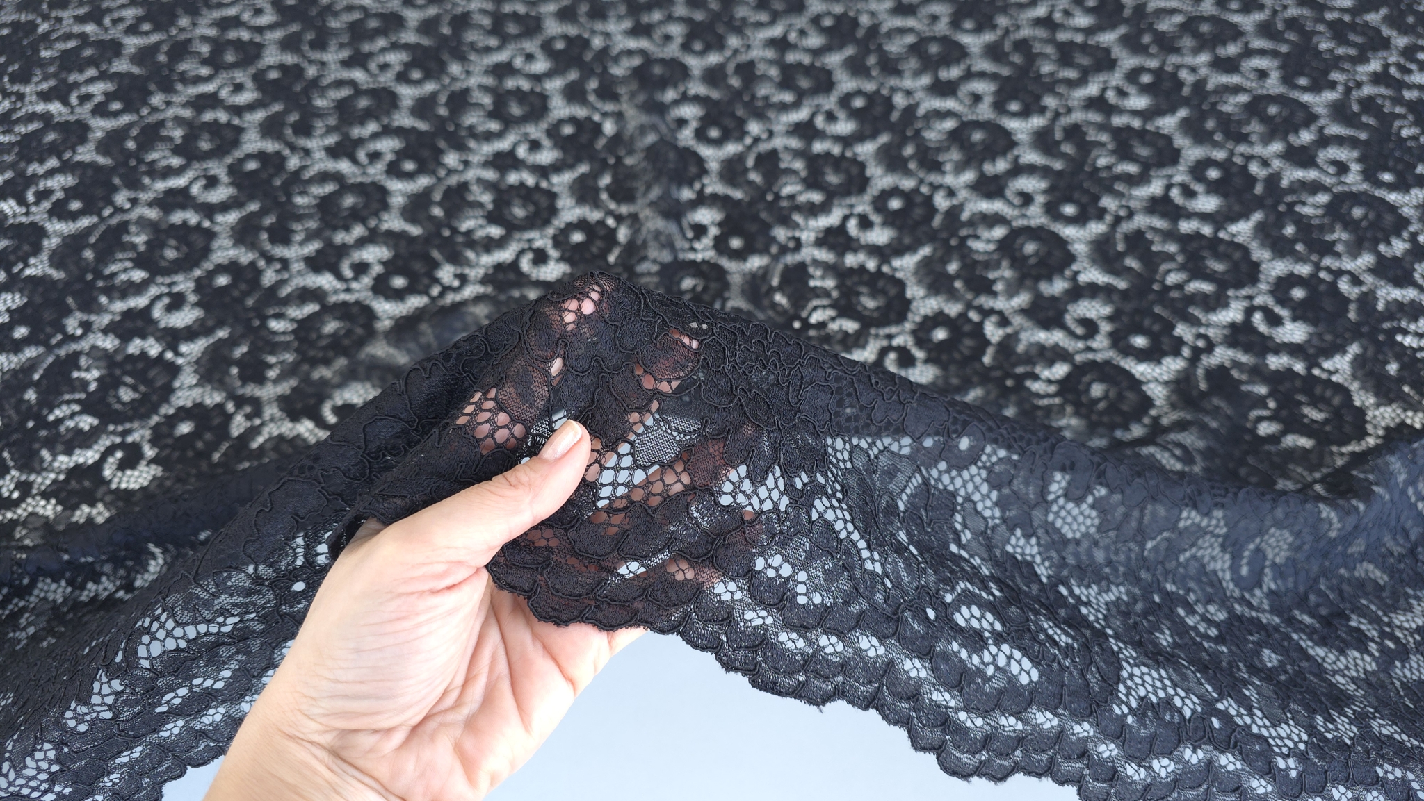 Mână care ridică dantelă neagră cu model floral evidențiind bordura festonată și transparența materialului