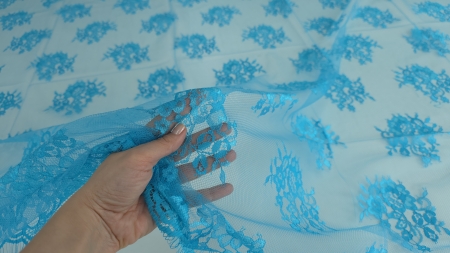 Mână care ridică dantela turquoise de pe suprafață cu buchete florale distribuite pe tull transparent