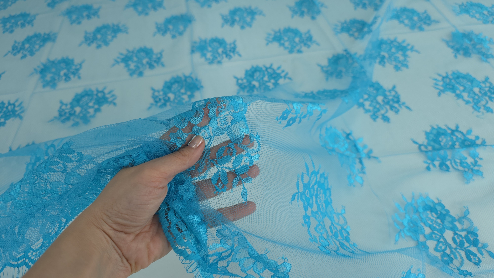 Mână care ridică dantela turquoise de pe suprafață cu buchete florale distribuite pe tull transparent