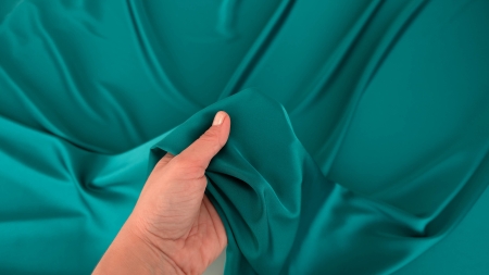 Mână care strânge material verde albastru profund, falduri moi ce se formează natural în jur