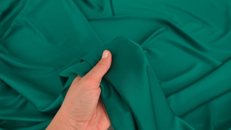 Mână care ține material verde smarald rulat în cornet între degete, detaliu textură fină și revenire elastică