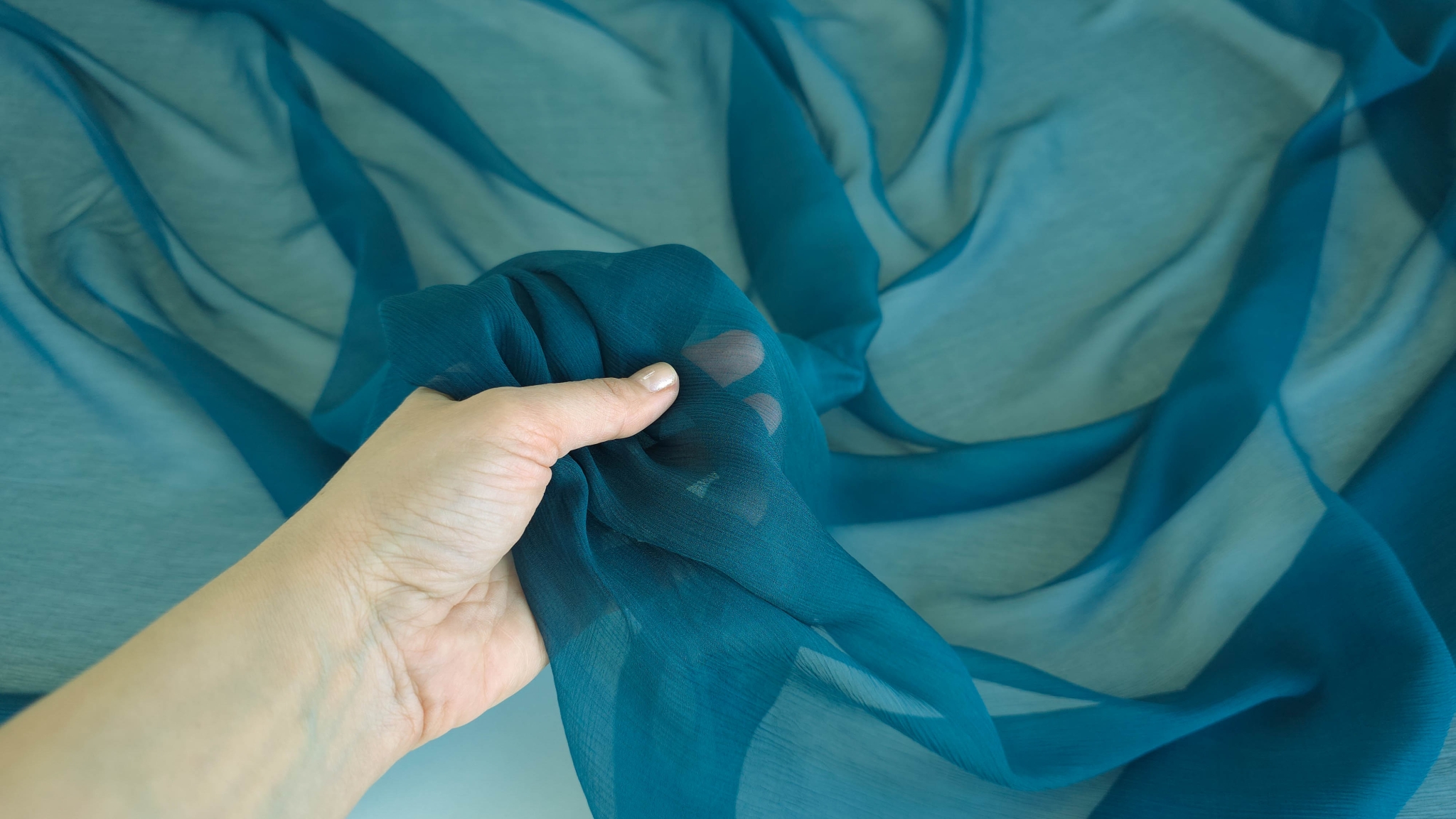 Mână ridicând muselină din mătase naturală albastru petrol arătând transparența completă
