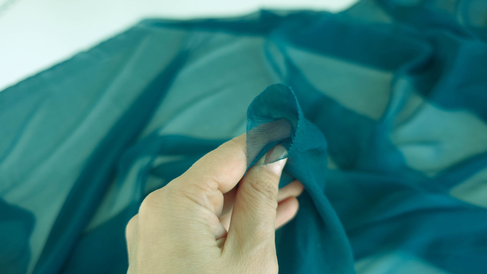 Mână ținând un colț de voal albastru petrol cu marginea tăiată vizibilă între degete