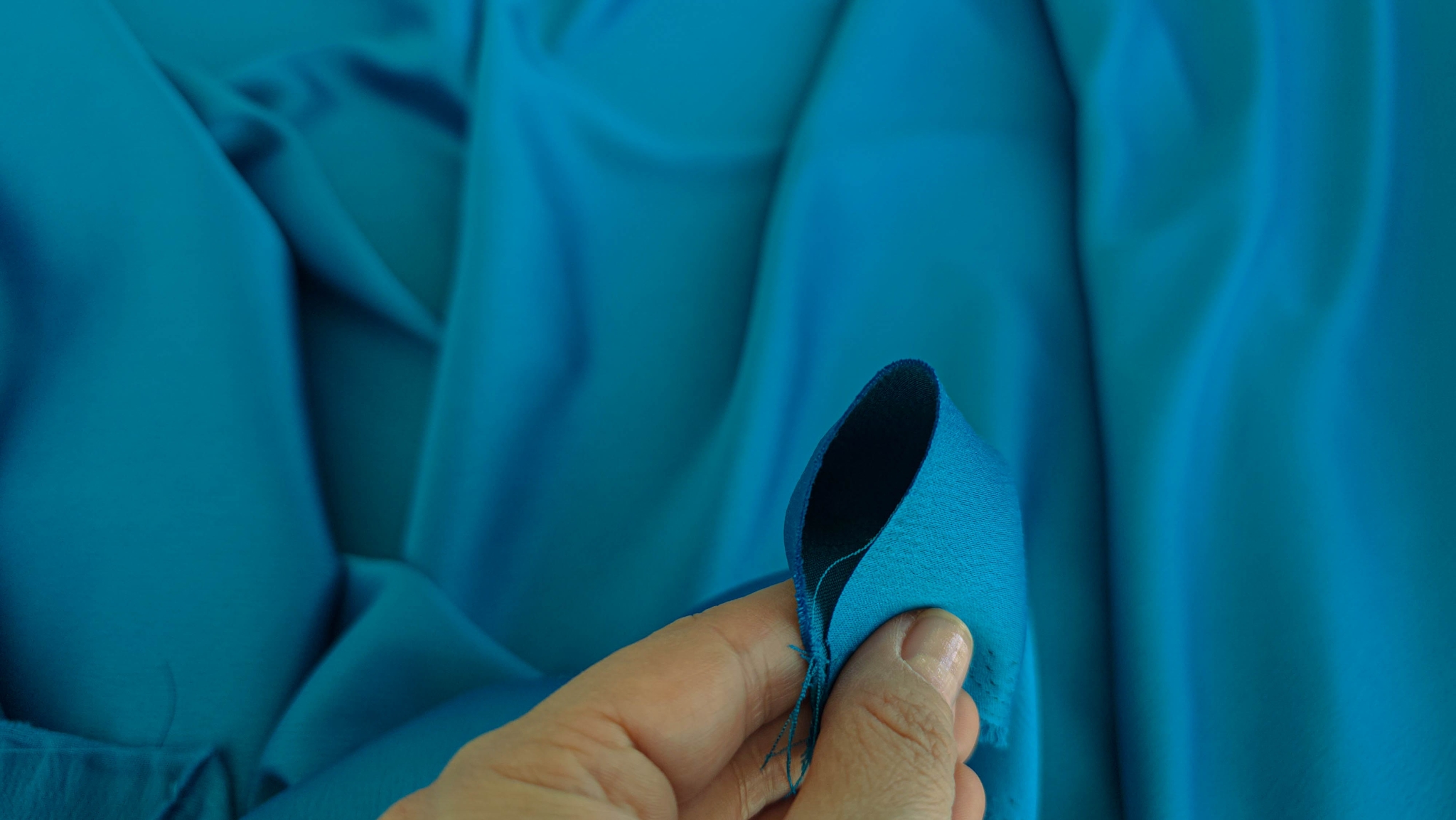 Mână ține marginea îndoită a taftei turquoise, contrast clar între față lucioasă și dos mat