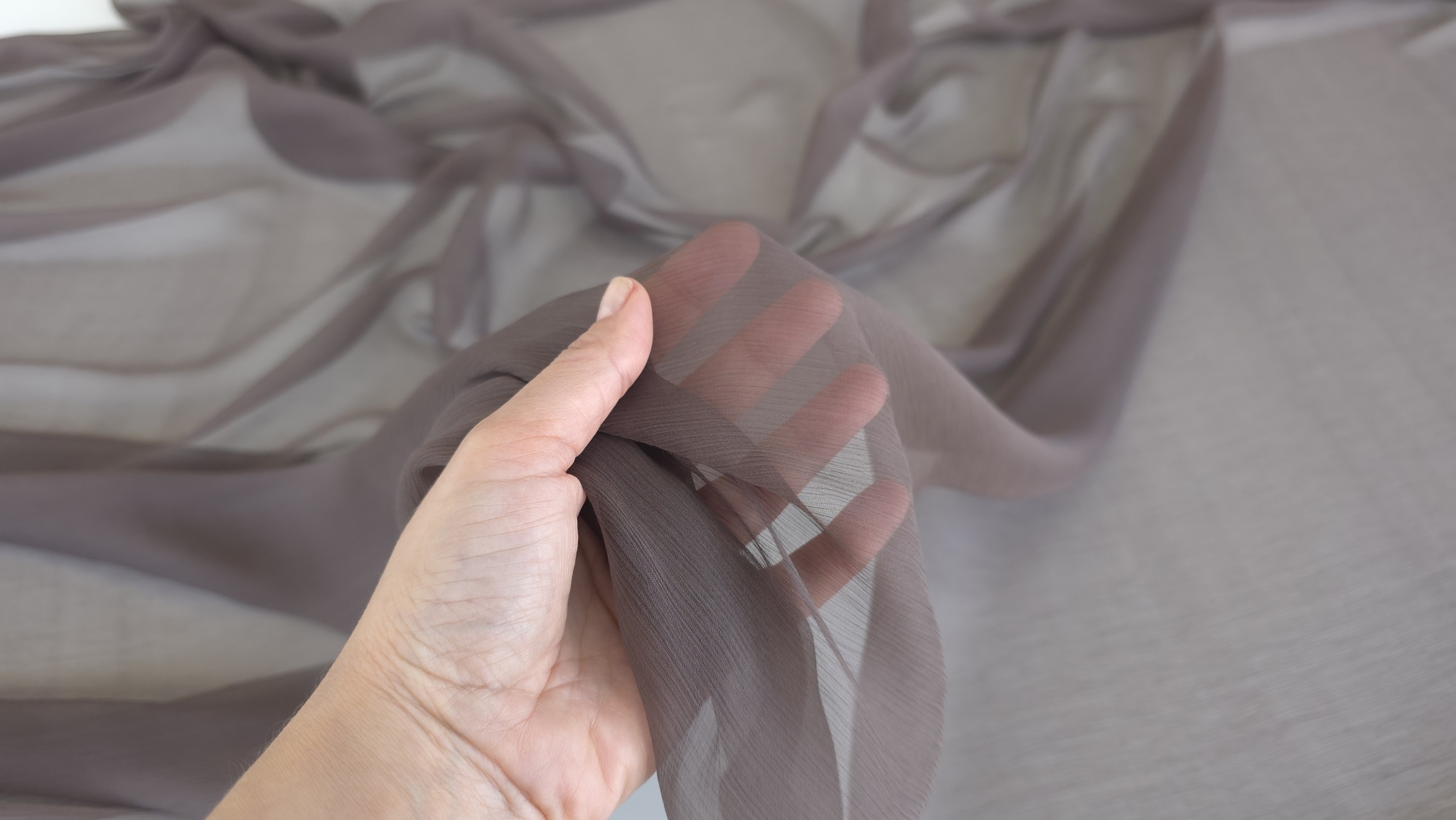 Materialul transparent ținut în palmă cu degetele vizibile prin țesătura creponată maro grej