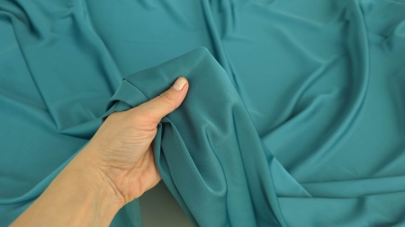Mână care ține material turquoise rulat în cornet mic între degete, detaliu textură fină și revenire elastică