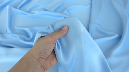 Mână ține material satinat sky blue în falduri libere, cadru larg