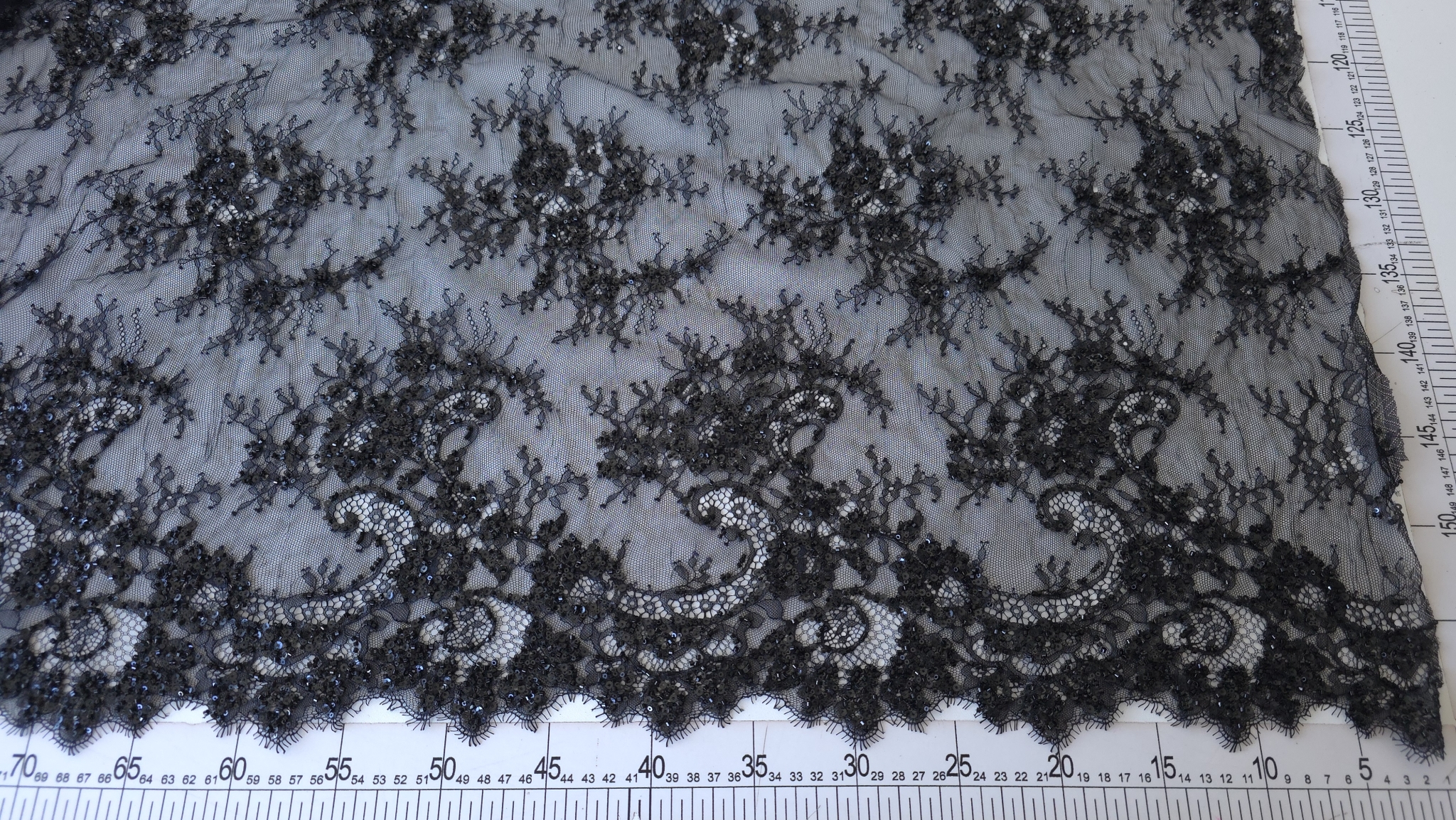 Dantela tip chantilly negru accesorizata, cod 60254, întinsă pe masă lângă riglă gradată