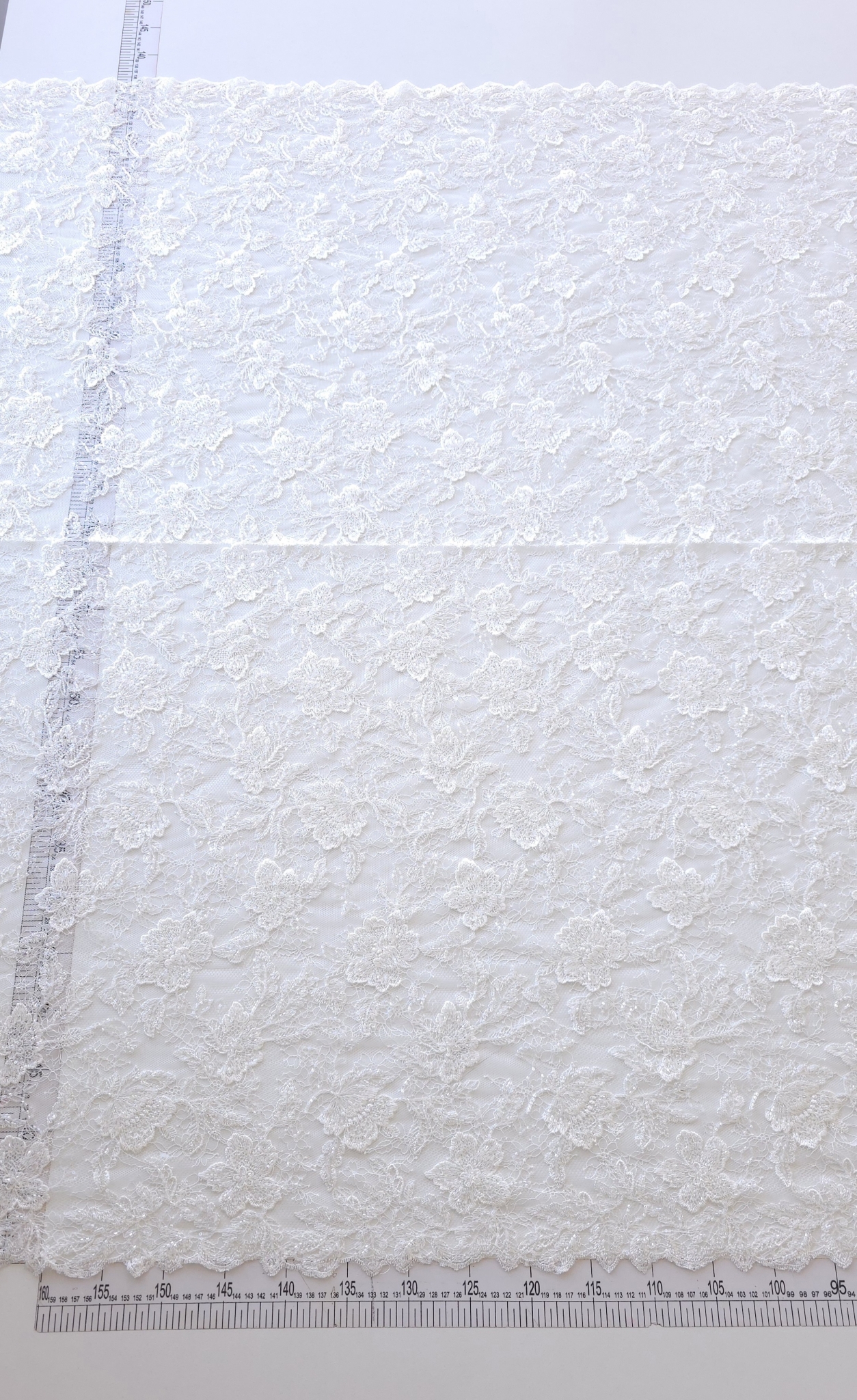 Dantela tip chantilly off white accesorizata cu margele întinsă pe masă albă lângă riglă
