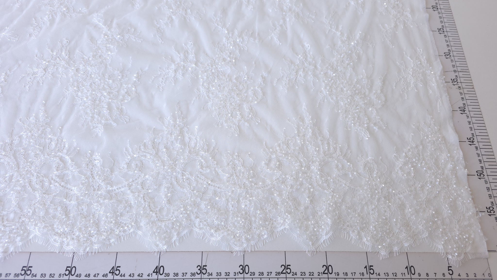 Dantela tip chantilly off white accesorizata cu paiete întinsă pe masă lângă riglă, cod 60260